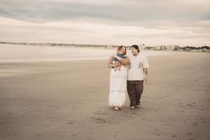 Family of three walking on a sandy beach, parents holding baby, with ocean and city skyline in background.