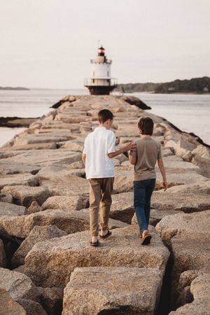 Two boys walk along a rocky jetty toward a lighthouse on the water at dusk.