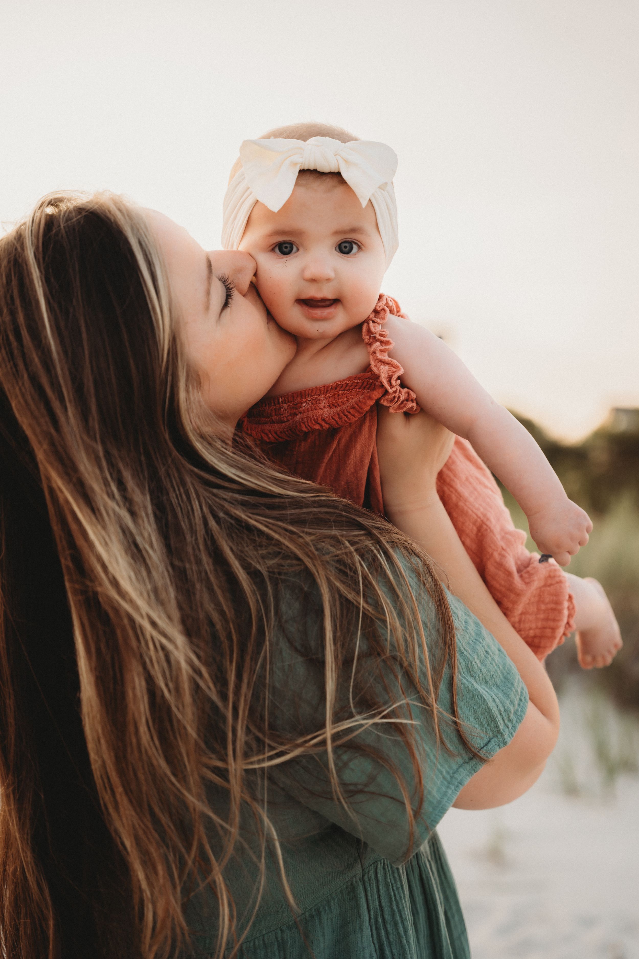 A mother with long hair kisses her baby girl in a pink bow headband outdoors at golden hour.