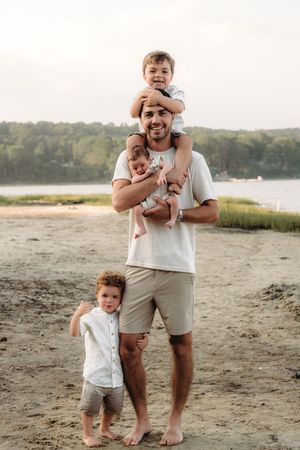 A smiling father stands on a sandy beach holding a newborn while two young boys climb on him, with trees and water behind.