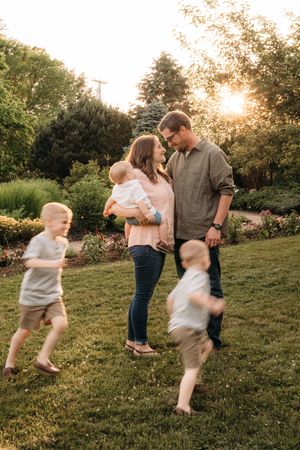 Family with two young boys running and parents holding baby at golden hour in lush garden.