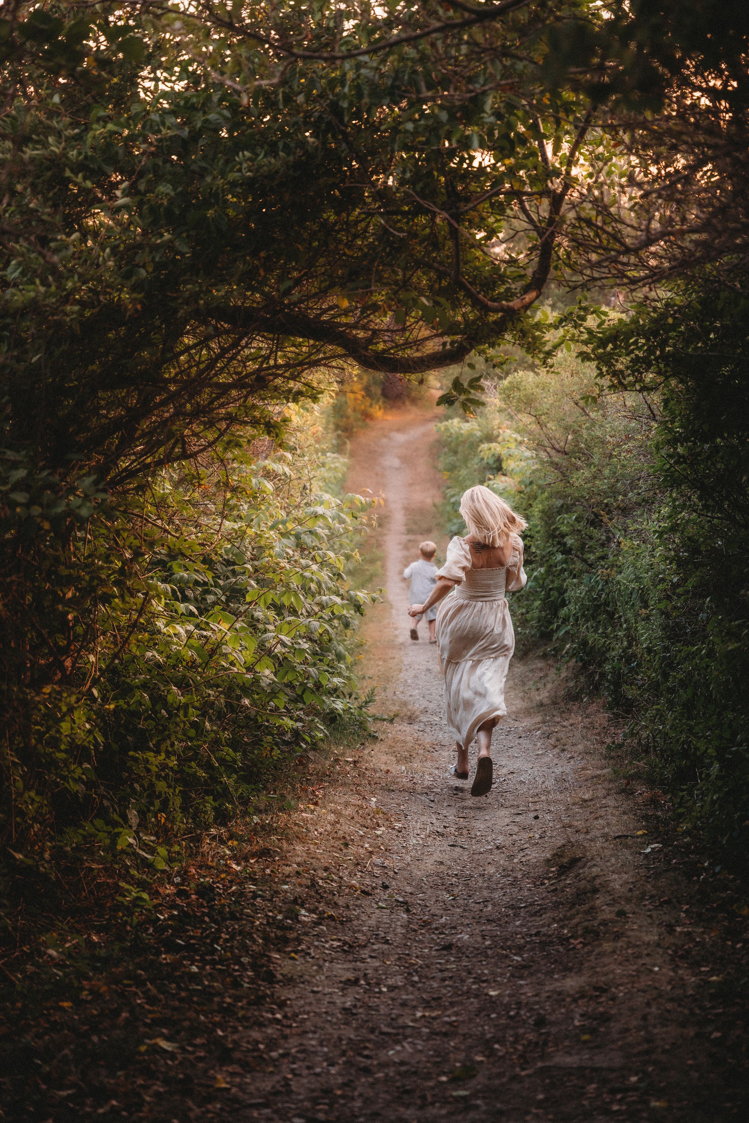 A blonde woman in a flowy white dress carries a child down a serene forest path surrounded by lush green trees.