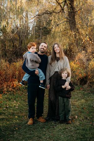 Family of four poses outdoors in autumn park, parents holding two young red-haired boys among colorful fall foliage.