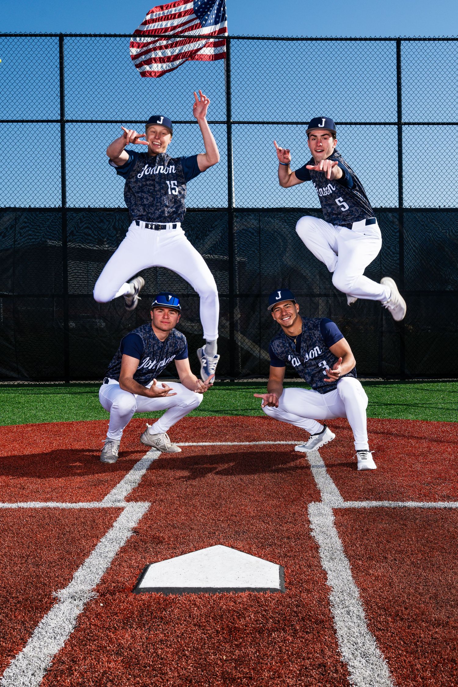 Four baseball players pose at home plate with an American flag, two jumping and two crouching, in navy uniforms.