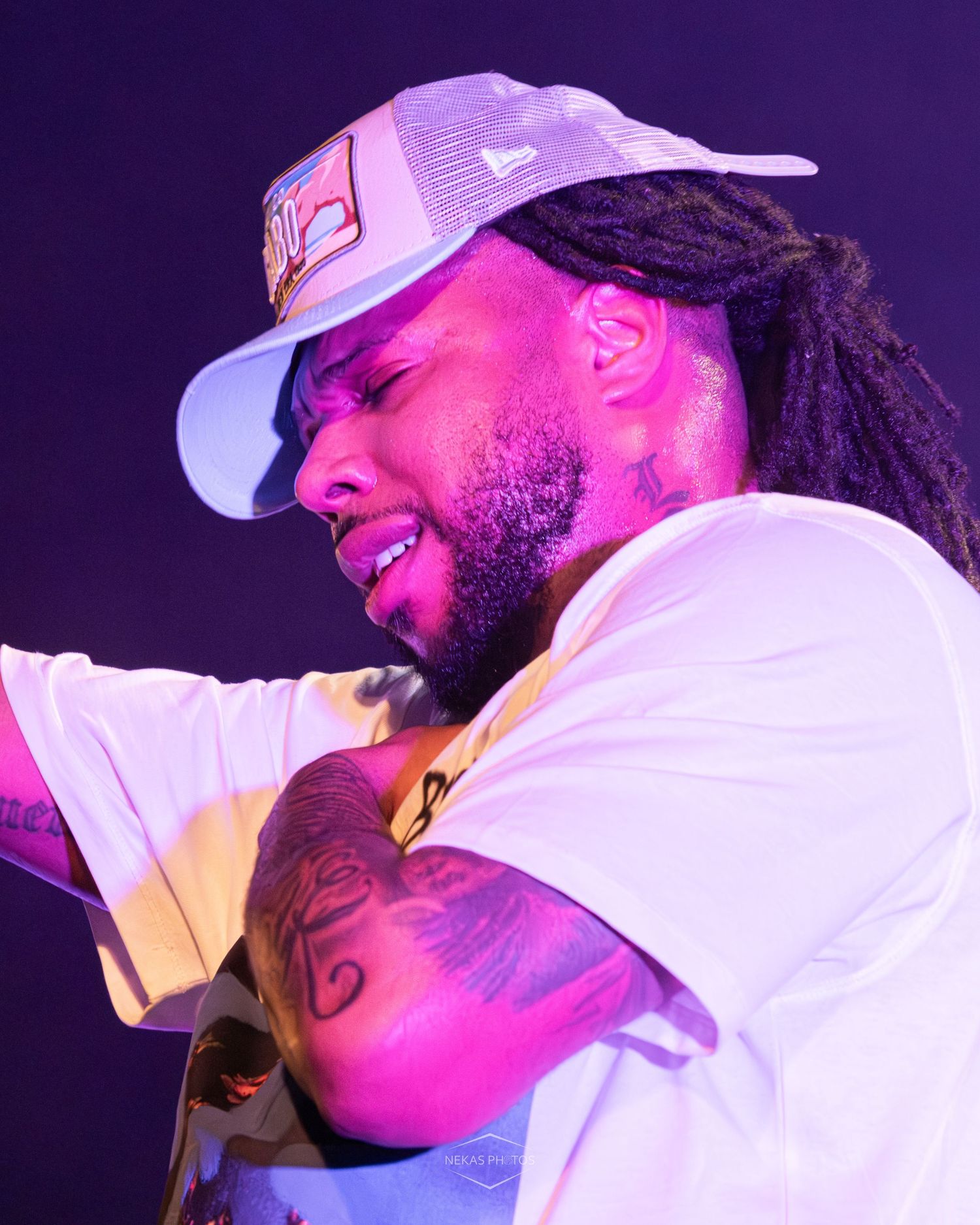 A tattooed man with dreadlocks and a white trucker hat performs on stage under pink and purple lights.