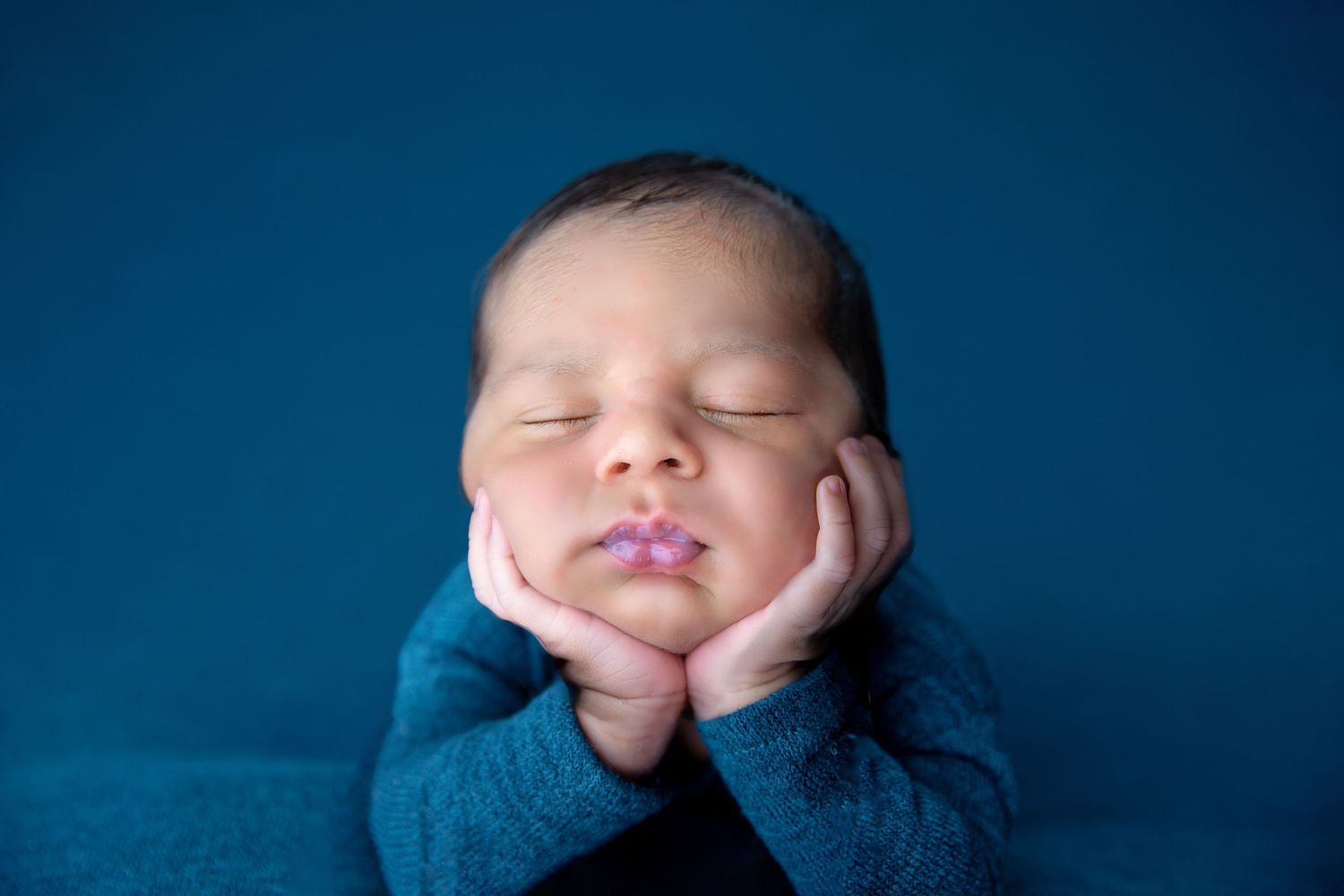 Peaceful newborn baby sleeping with hands on cheeks, wrapped in teal knit blanket against blue background.
