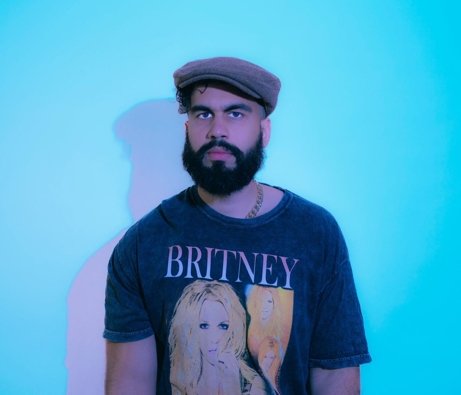 Bearded man wearing a Britney Spears graphic tee and flat cap poses against a teal background.