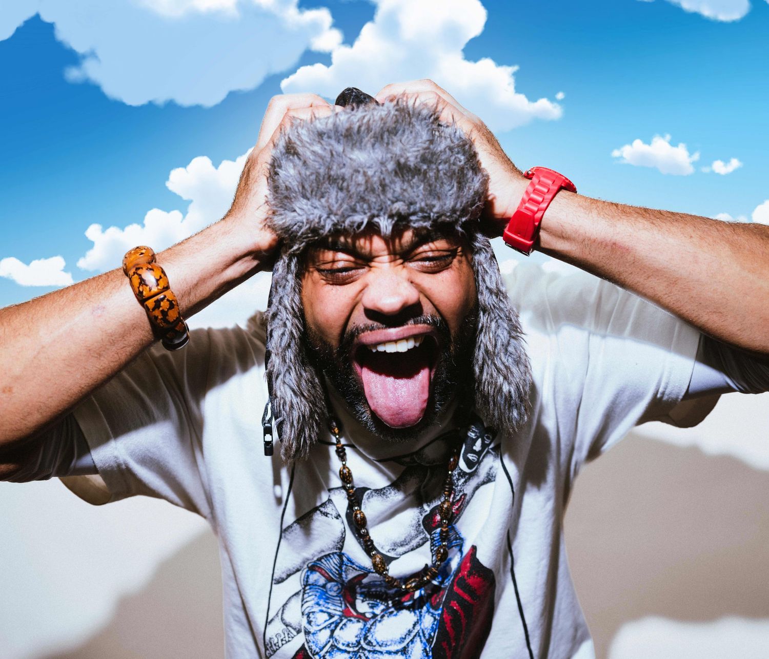 Man wearing fur trapper hat sticking tongue out with hands on head against blue sky background.