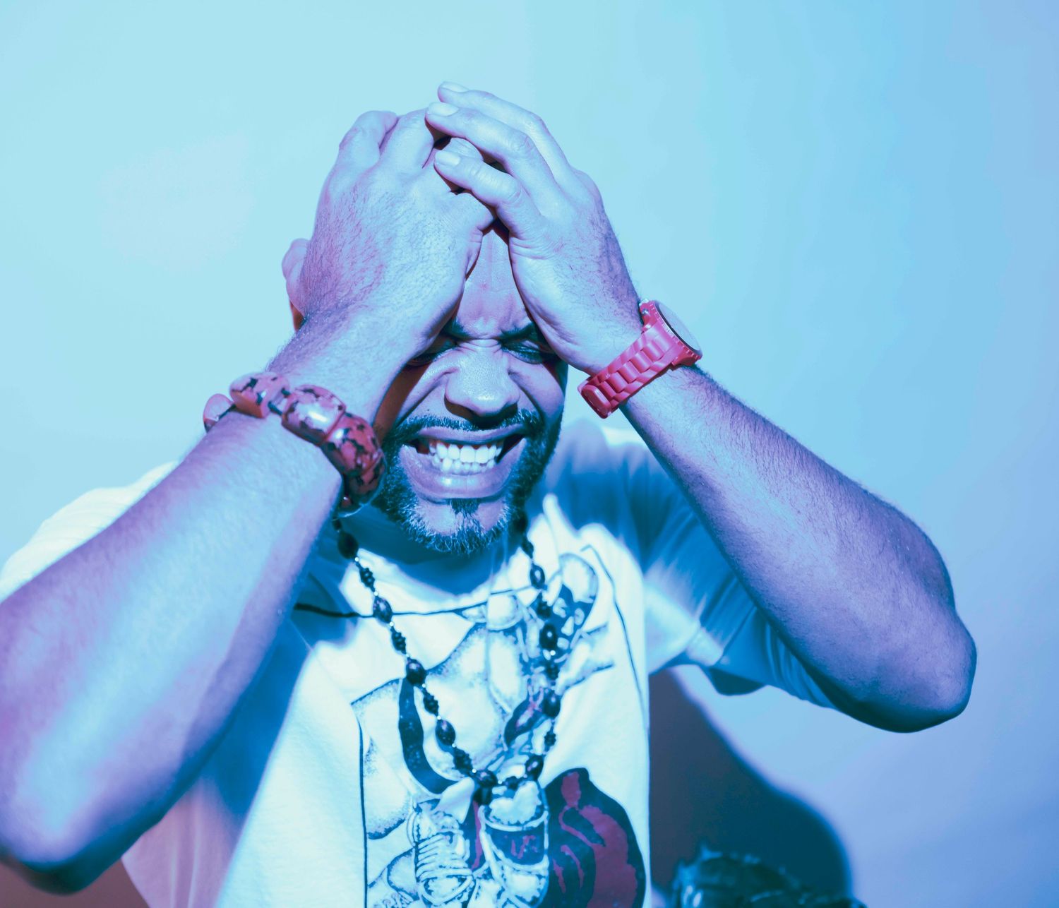 Man in white graphic tee and red bracelets holds head in hands, laughing under blue-purple lighting.