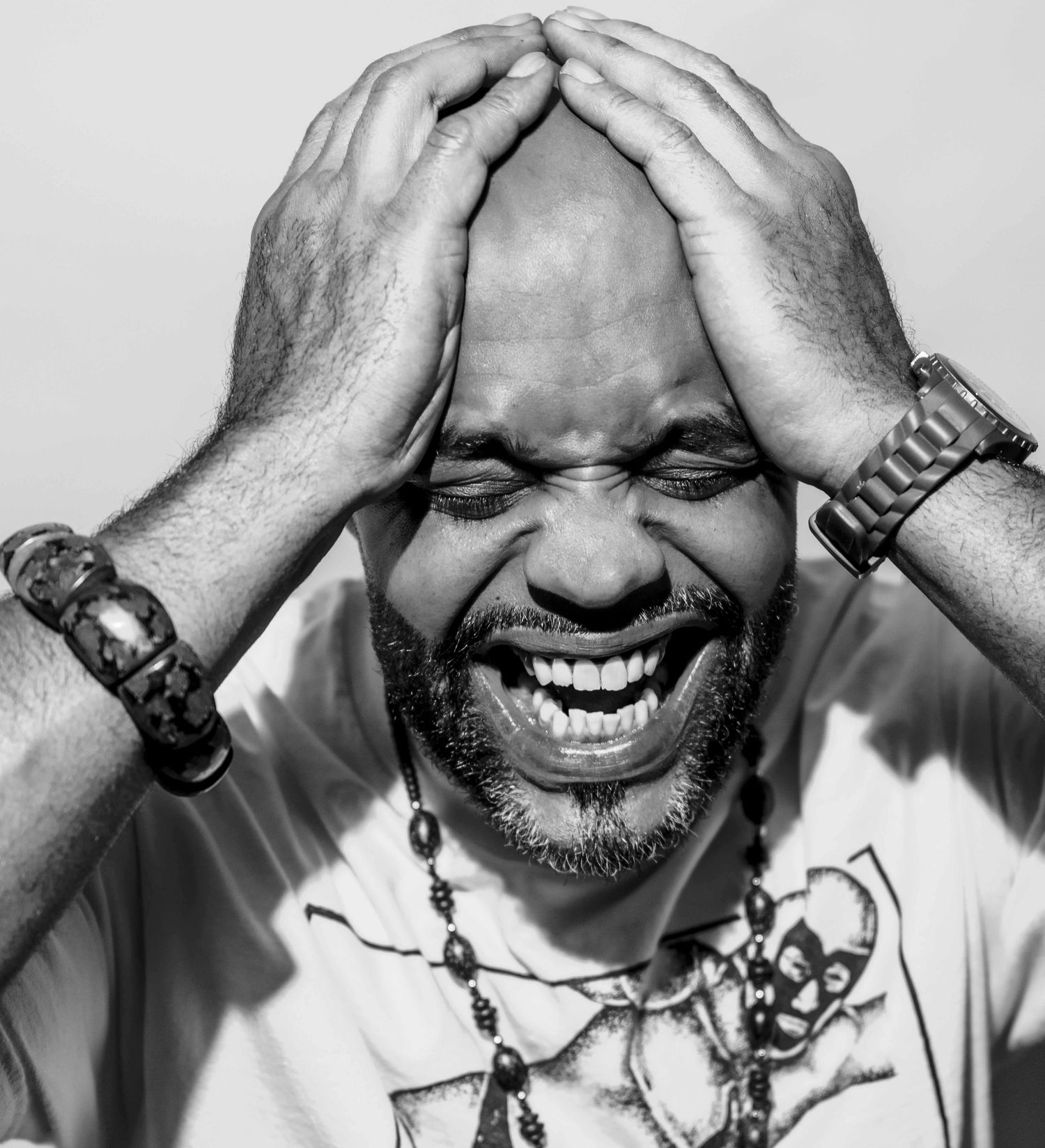 Black and white photo of a laughing bald man with beard, hands on head, wearing bracelets and necklace.