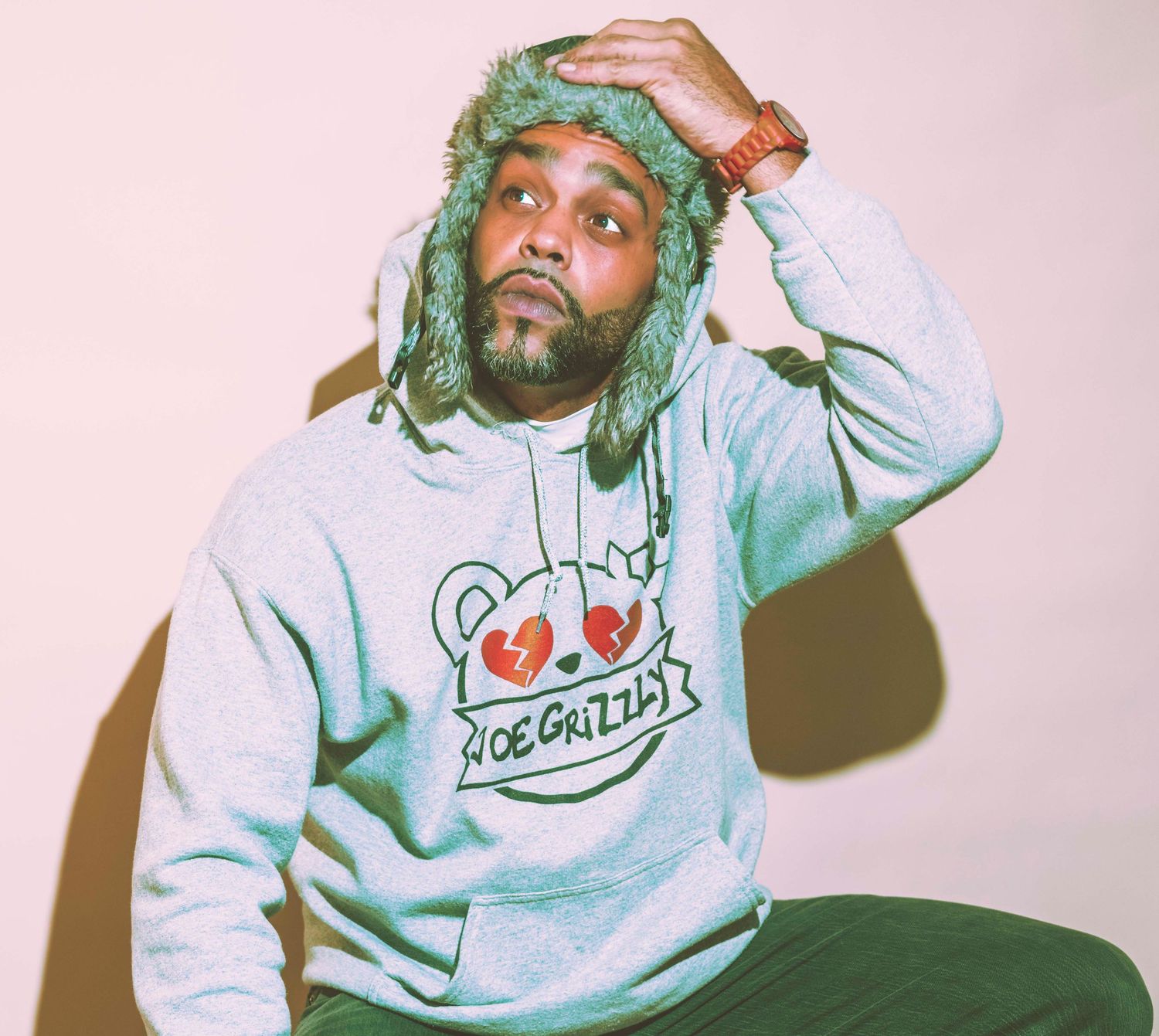 Man in gray Joe Grizzly hoodie and green fuzzy hat poses with hand on head against pink background.