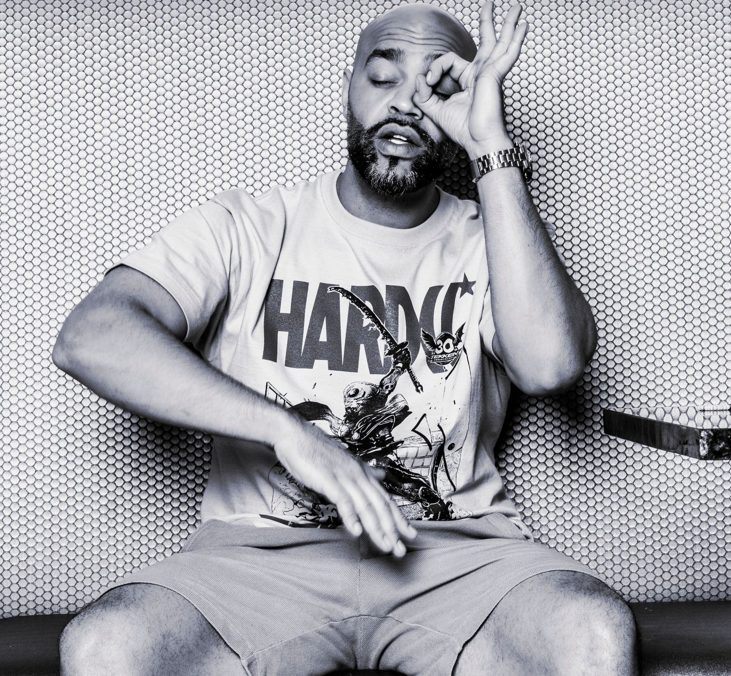 Man in HARDCO t-shirt and durag sits cross-legged making OK gesture, black and white portrait photo.