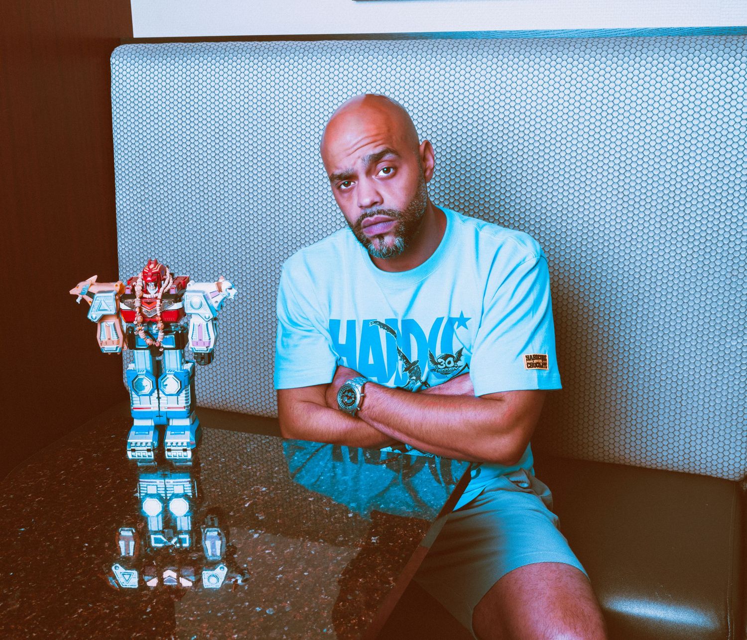 Bald man in light blue graphic tee sits with arms crossed next to a Voltron robot toy on a table.