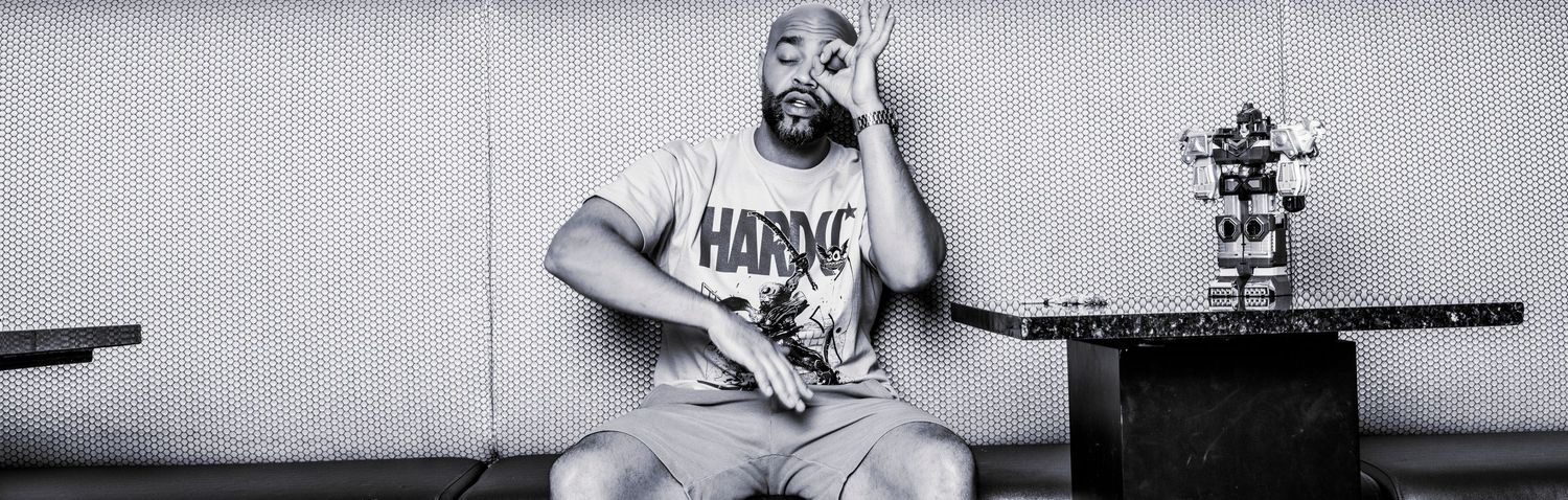 Black and white photo of a bearded man in a Hardee's shirt sitting casually beside a small robot figurine on a box.