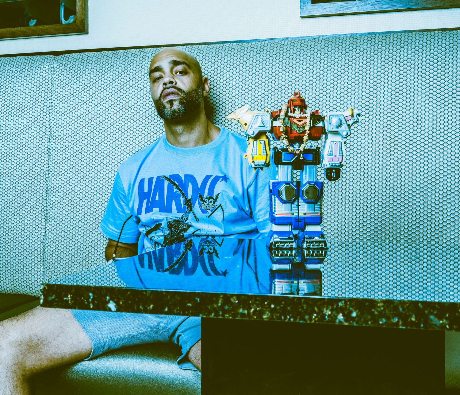 A man in a blue Hardcore graphic tee sits beside a colorful robot toy against a dotted blue wall.