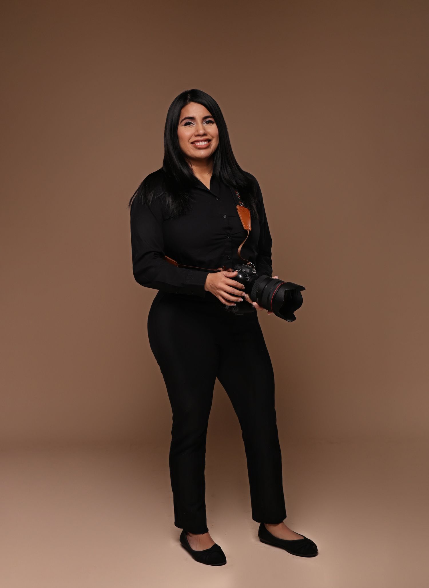 Woman in black outfit holding a professional camera, smiling, posing against a warm brown studio background.