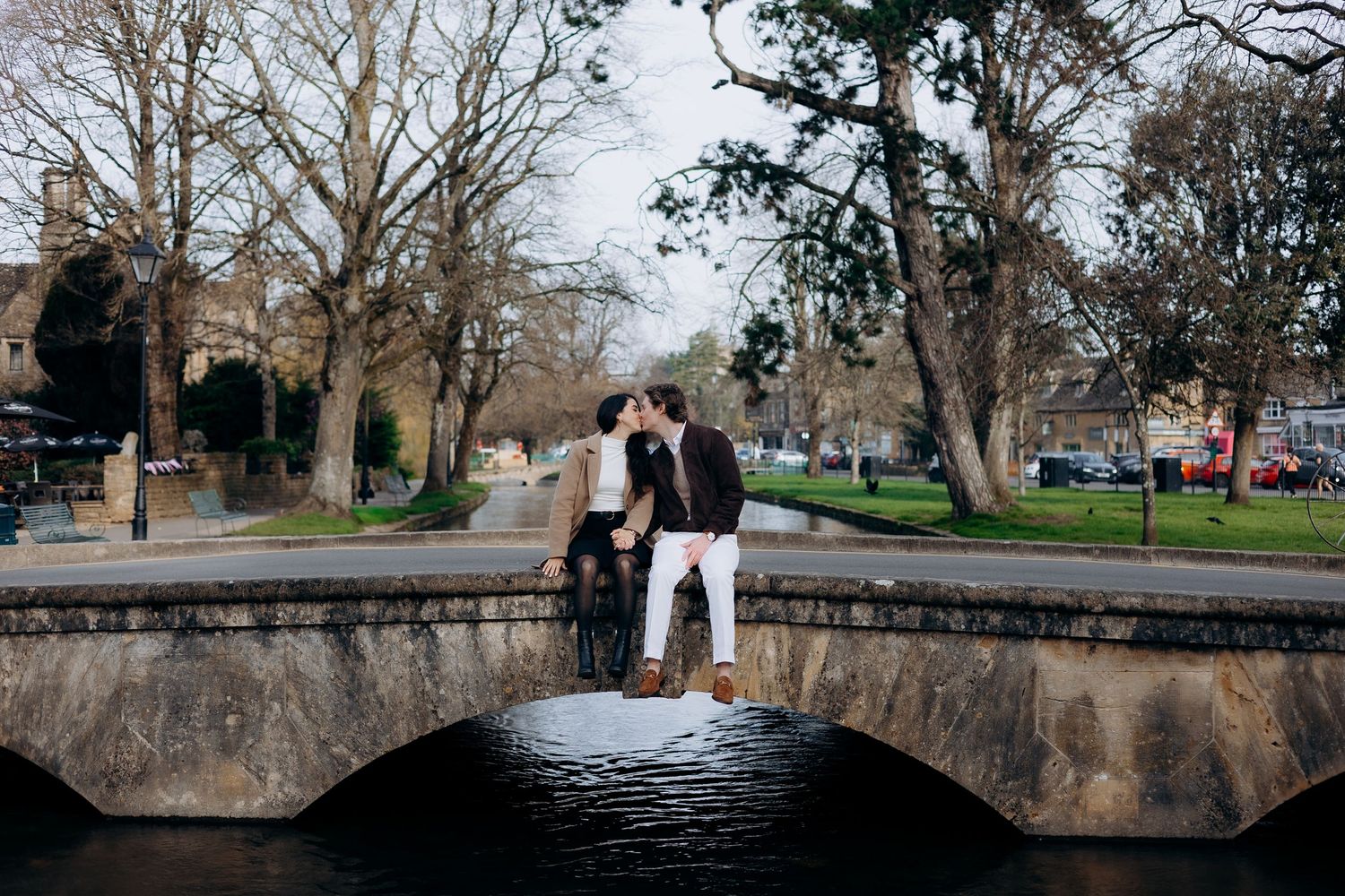 Couple sharing a romantic kiss at bourton on the water over a calm river surrounded by bare trees in a scenic park.