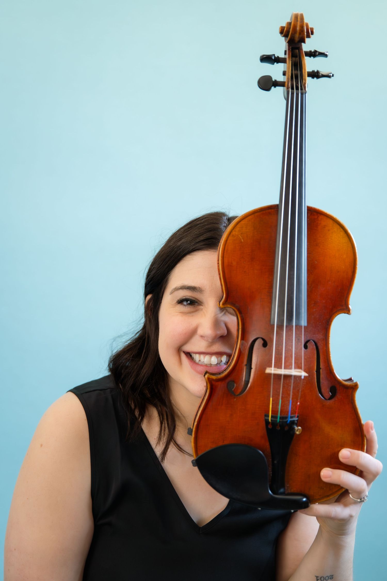 Smiling woman in black dress holding a violin against a light blue background.