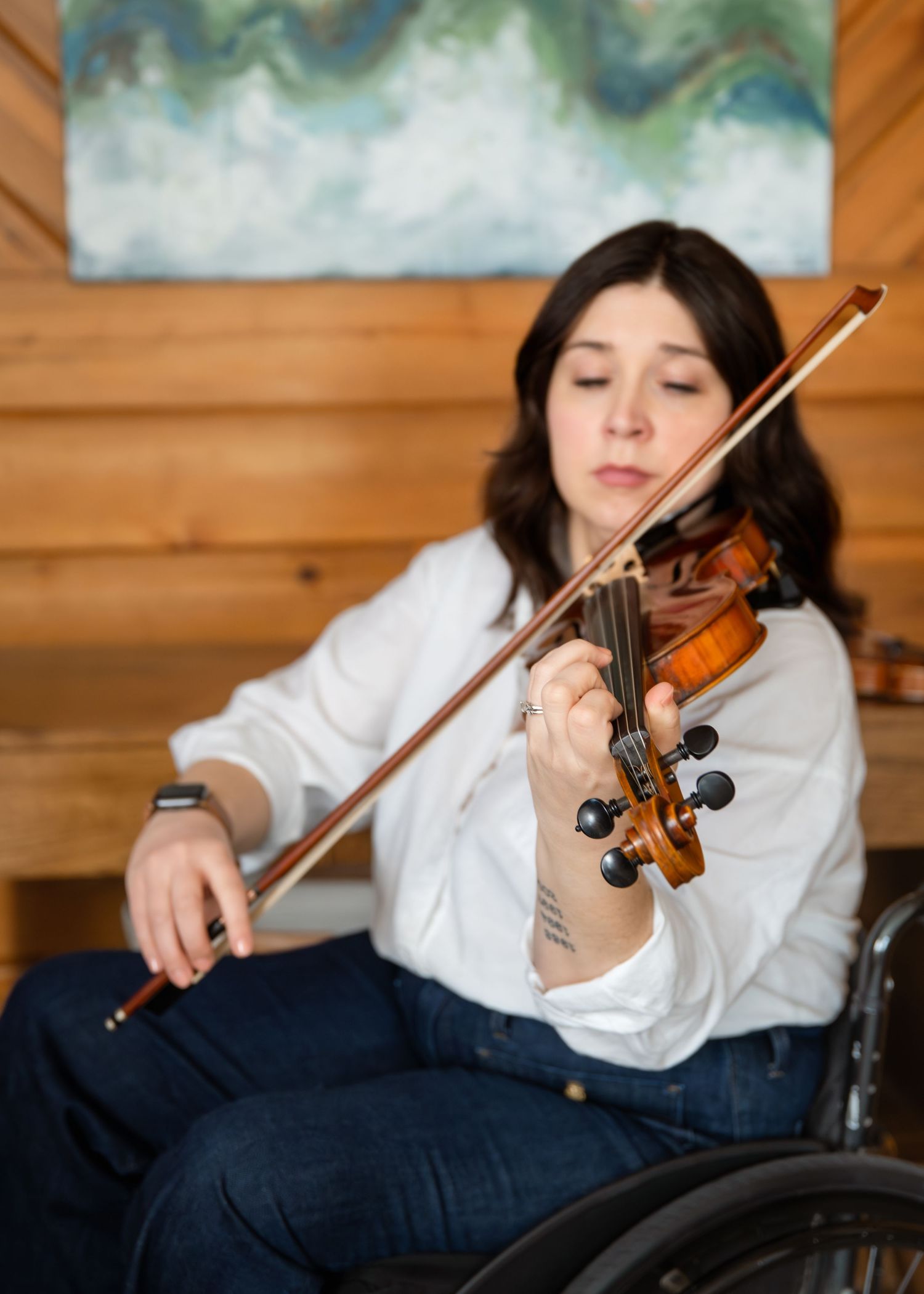 Woman in wheelchair playing violin indoors, wearing white shirt and jeans, focused on music near wooden wall.