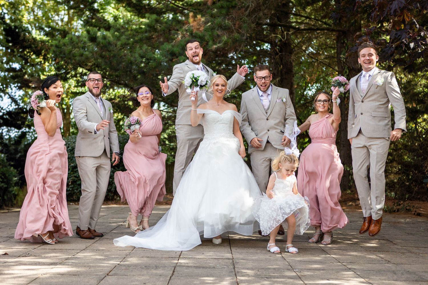 Large wedding party celebrates joyfully on a waterfront dock with colorful dresses and bouquets raised high.