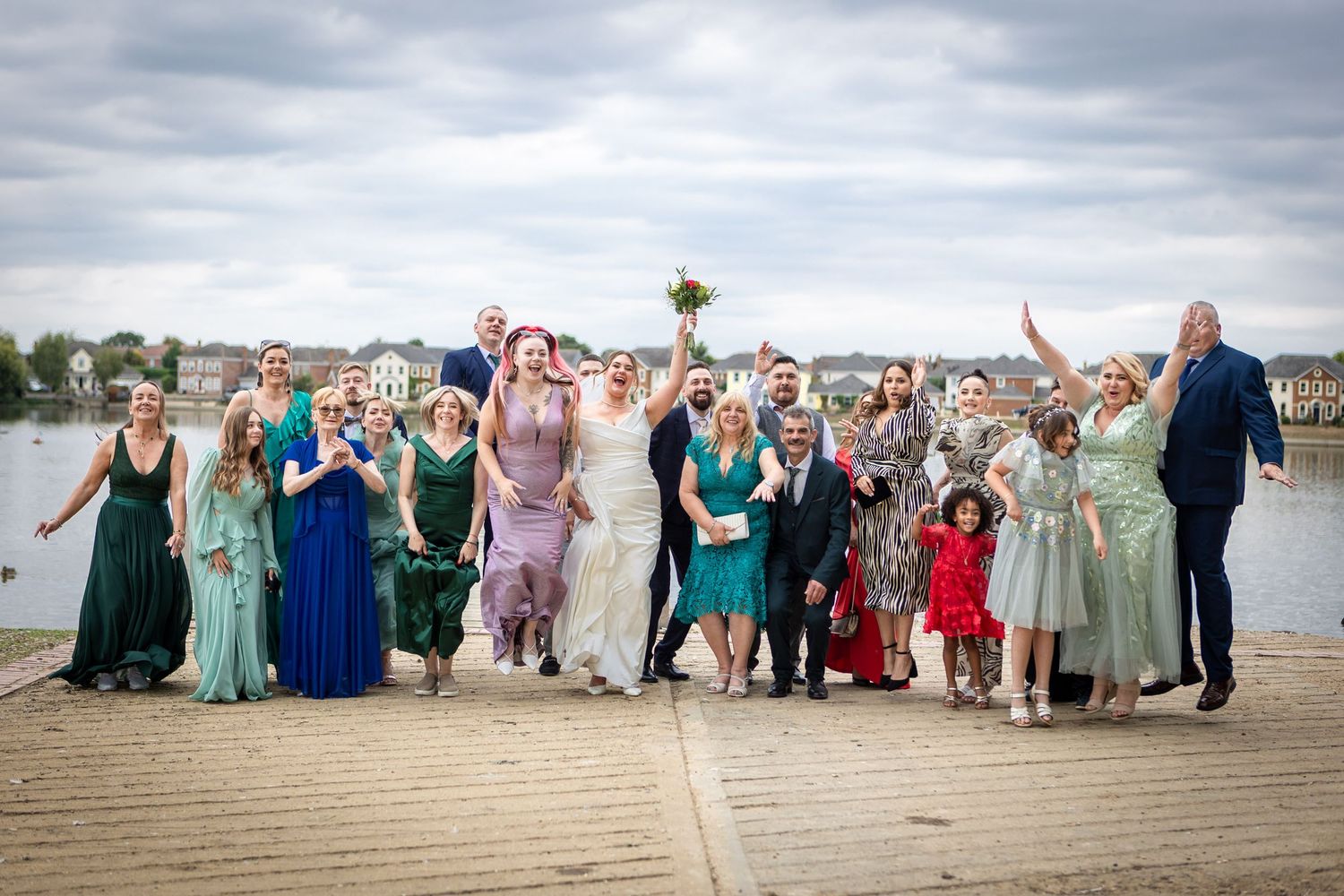 Large wedding party celebrates joyfully on a waterfront dock with colorful dresses and bouquets raised high.