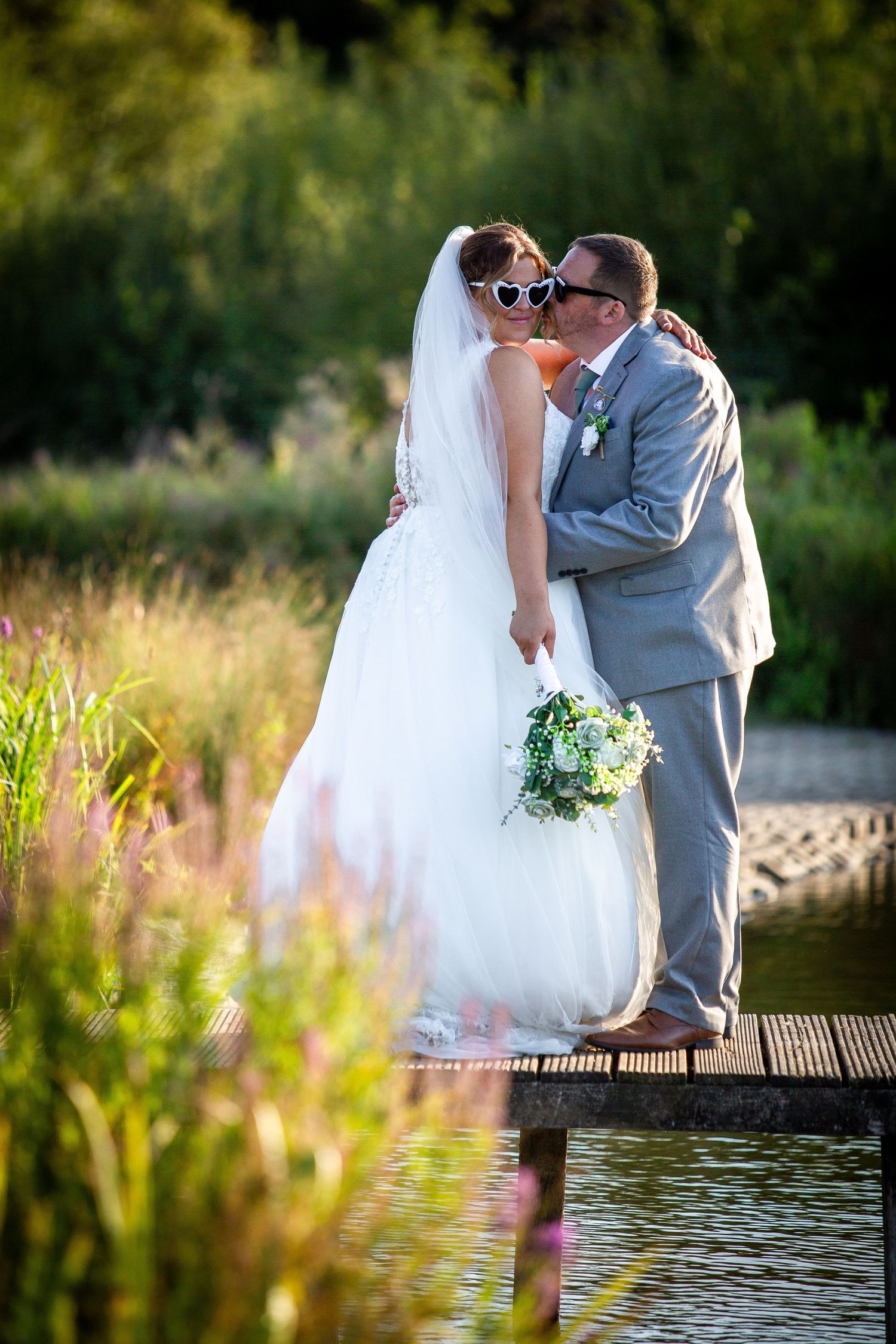 Large wedding party celebrates joyfully on a waterfront dock with colorful dresses and bouquets raised high.
