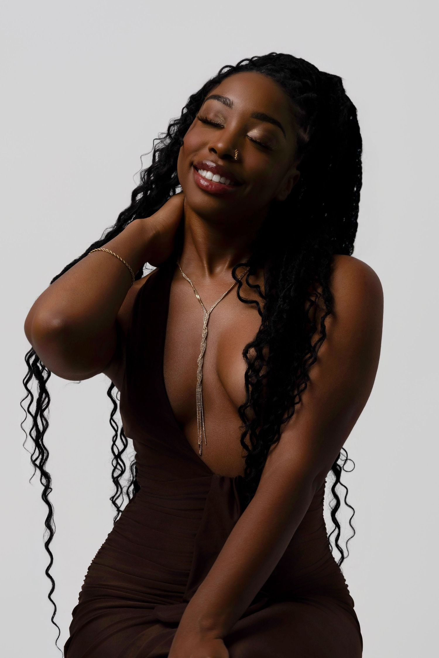 Smiling Black woman with long braided curly hair wearing a brown dress and gold necklace posing against a gray background.
