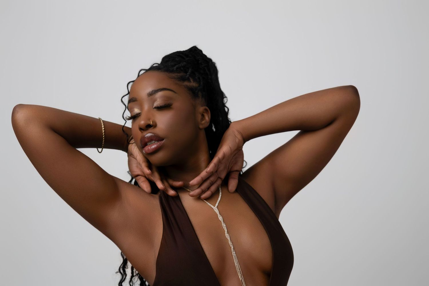 Young Black woman with braids poses gracefully, hands behind neck, wearing brown top and body chain jewelry.