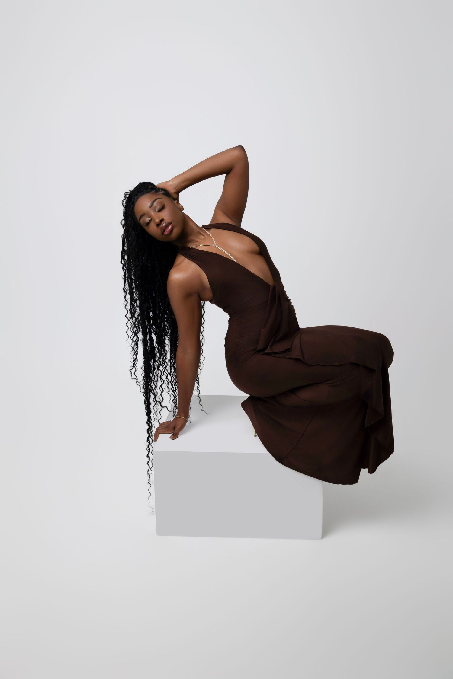A Black woman in a brown halter dress poses gracefully on a white cube, with long curly hair, against a light background.