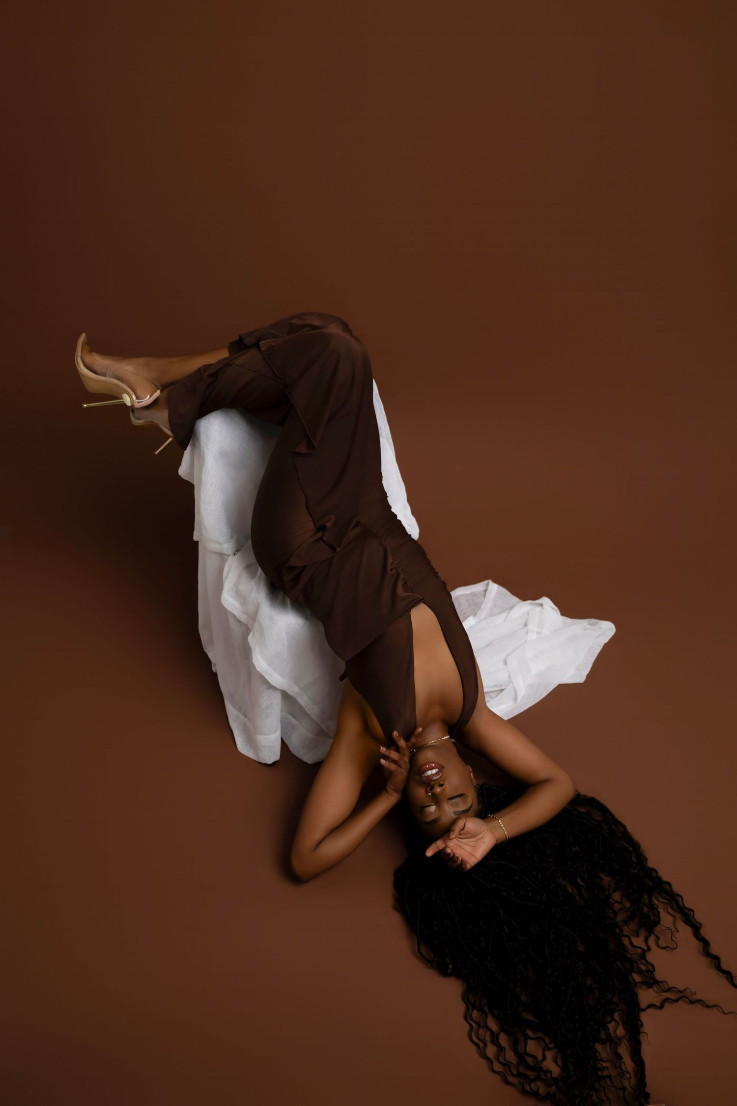 A Black woman in brown trousers and white shirt lies on a brown studio floor, legs raised, wearing gold heels.