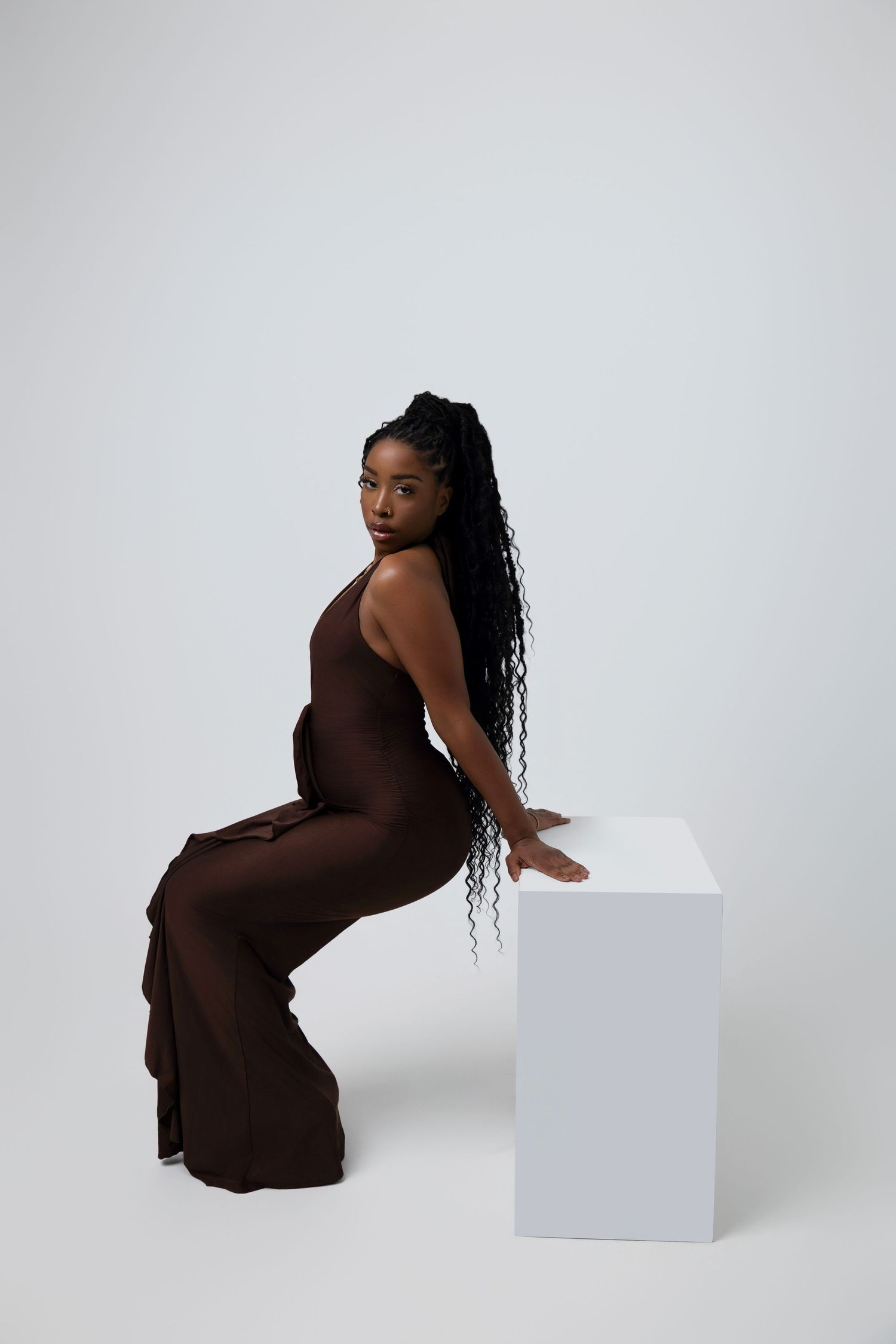 A Black woman with long braids sits elegantly on a white cube, wearing a brown wide-leg jumpsuit against a light background.