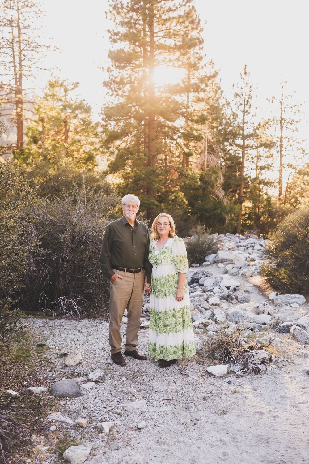 Older couple standing on rocky path surrounded by tall pine trees with golden sunlight streaming through.