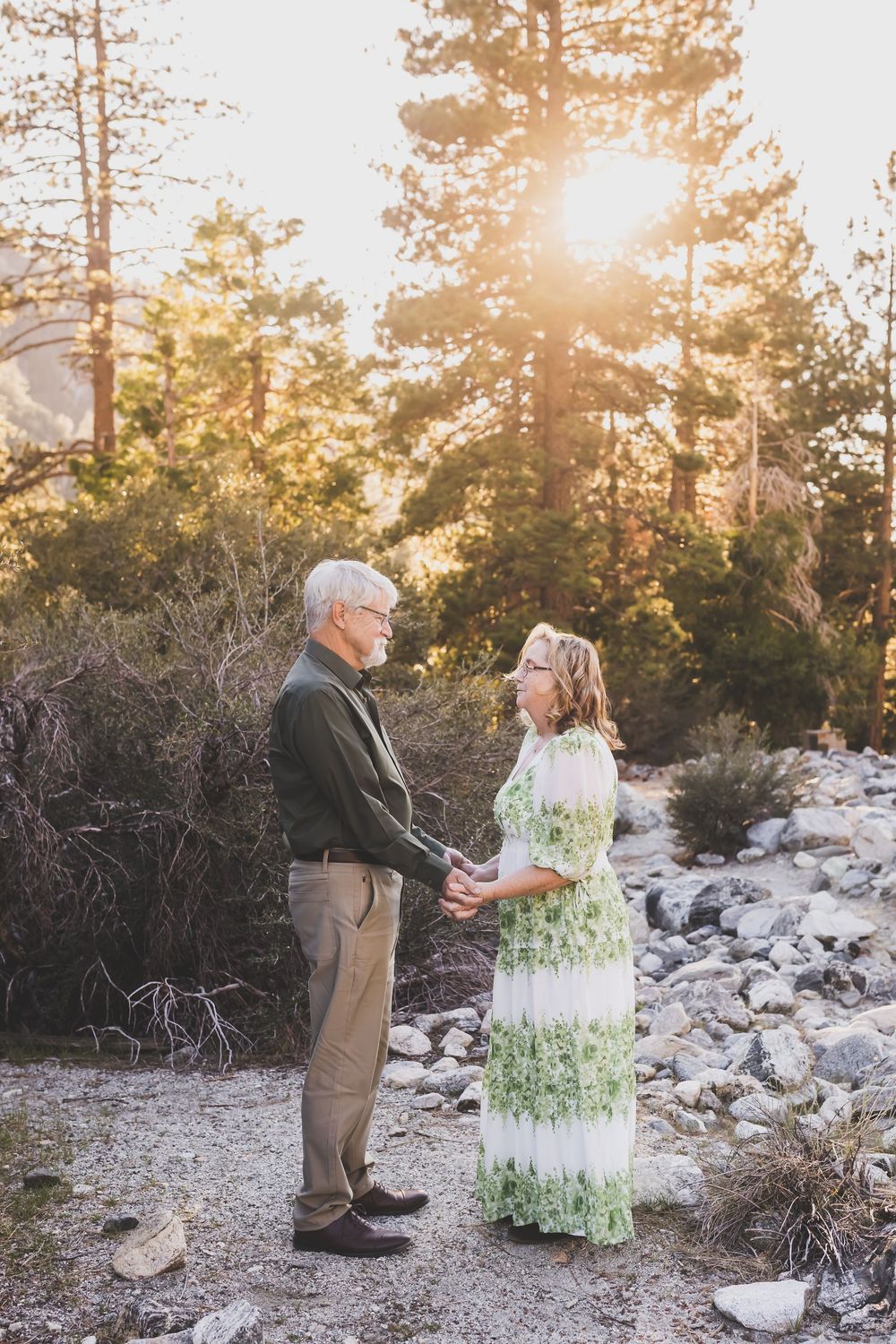 Older couple holding hands and gazing at each other in a sunlit forest setting with pine trees and rocks.