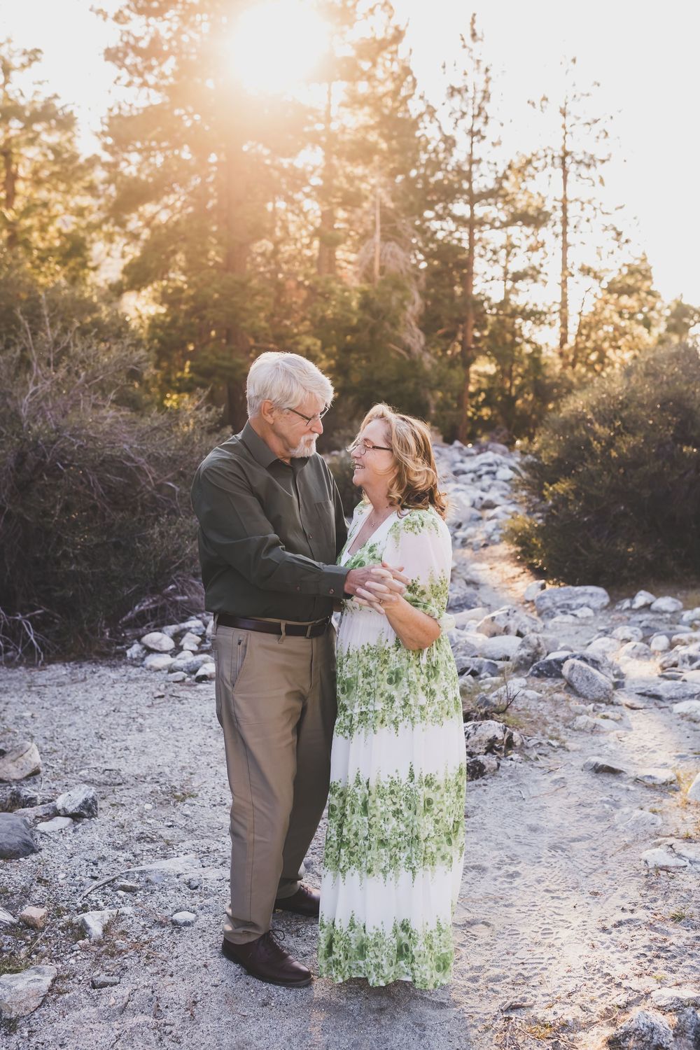 Elderly couple embracing outdoors at golden hour, surrounded by rocks and trees in a serene natural setting.