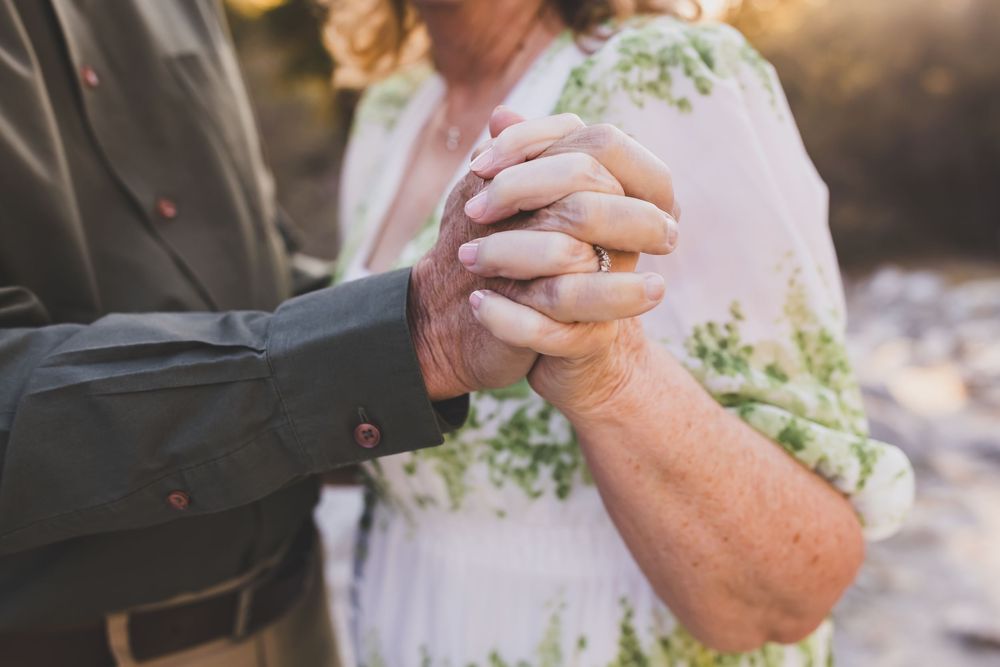 Elderly couple holding hands tenderly outdoors, woman in floral dress and man in olive jacket, symbolizing lasting love.