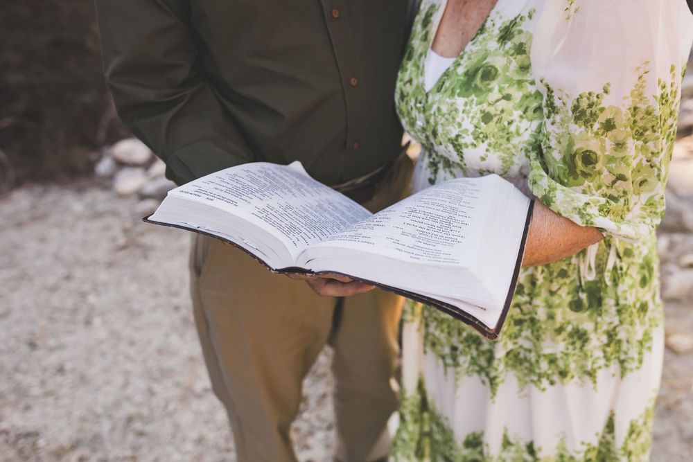 Couple holding open Bible during outdoor wedding ceremony, woman in floral green dress, man in olive jacket.