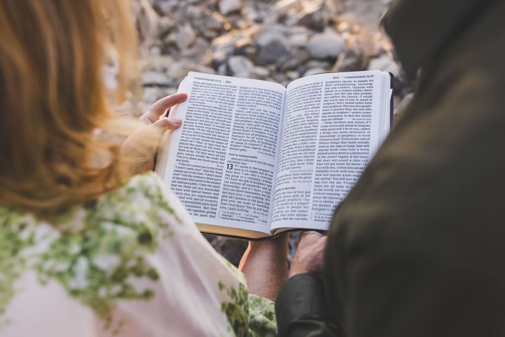 Couple reading an open Bible together outdoors, with woman holding flowers in a romantic setting.