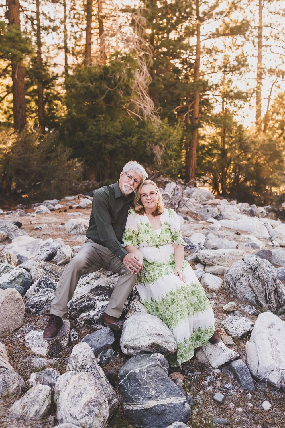 Mature couple sitting together on rocky terrain surrounded by pine trees at golden hour.