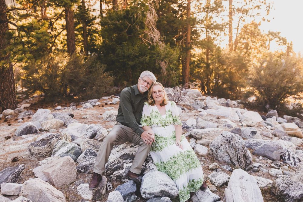 Couple sitting on rocks in a sunlit forest, man in gray embracing woman in green floral dress, golden hour photography.