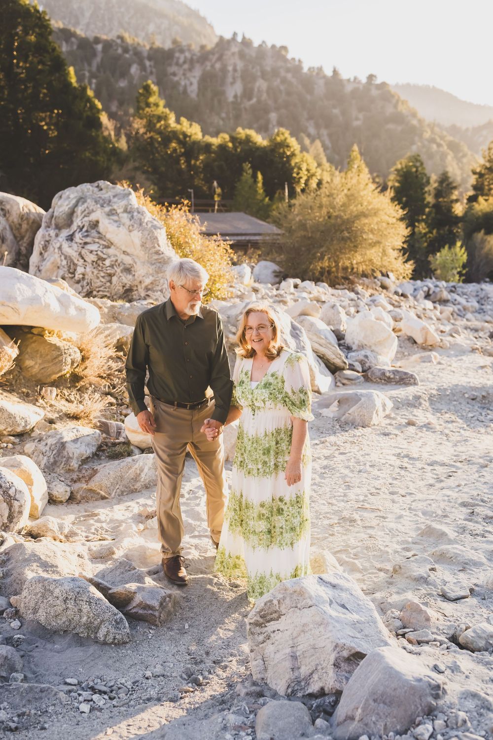Elderly couple holding hands walking through rocky mountain landscape at golden hour sunset.