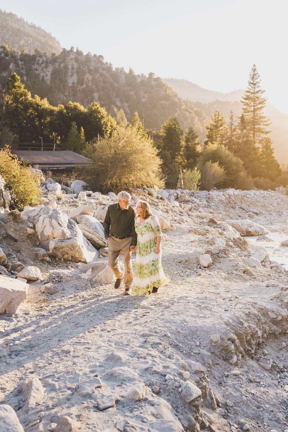 Senior couple walking hand in hand over rocky landscape with mountain and pine trees at sunset.