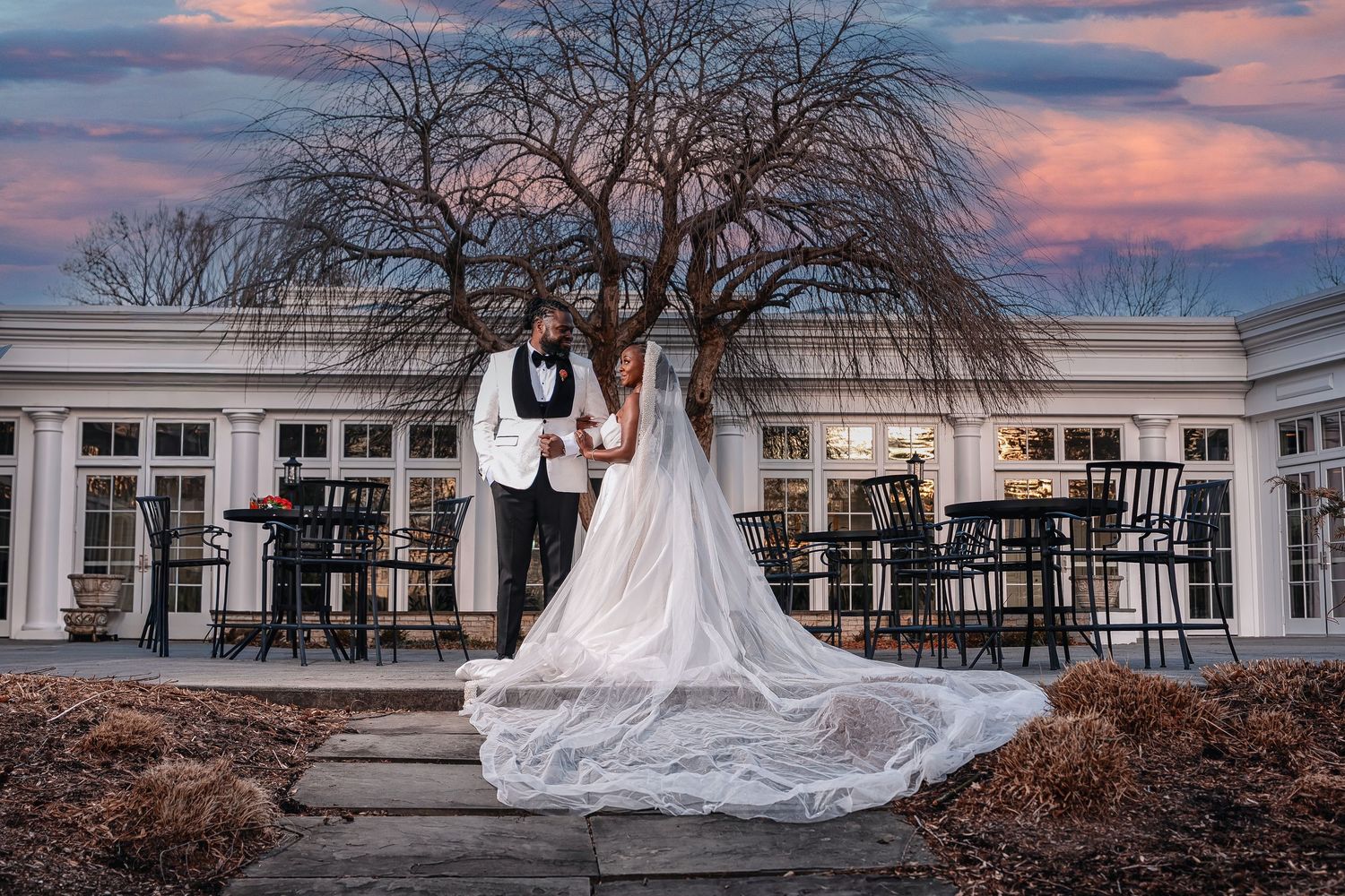 Bride and groom stand together before a white venue at dusk, her lace gown trailing elegantly on stone steps. Deerfield Golf Club Newark De