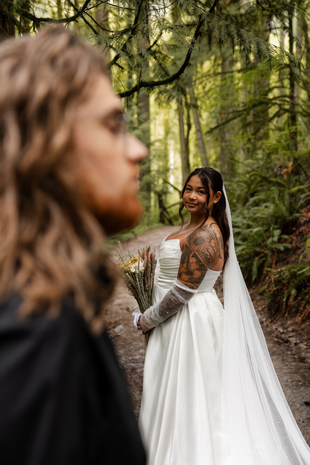 Tattooed bride in white gown holds bouquet on forest path, smiling at groom in foreground.