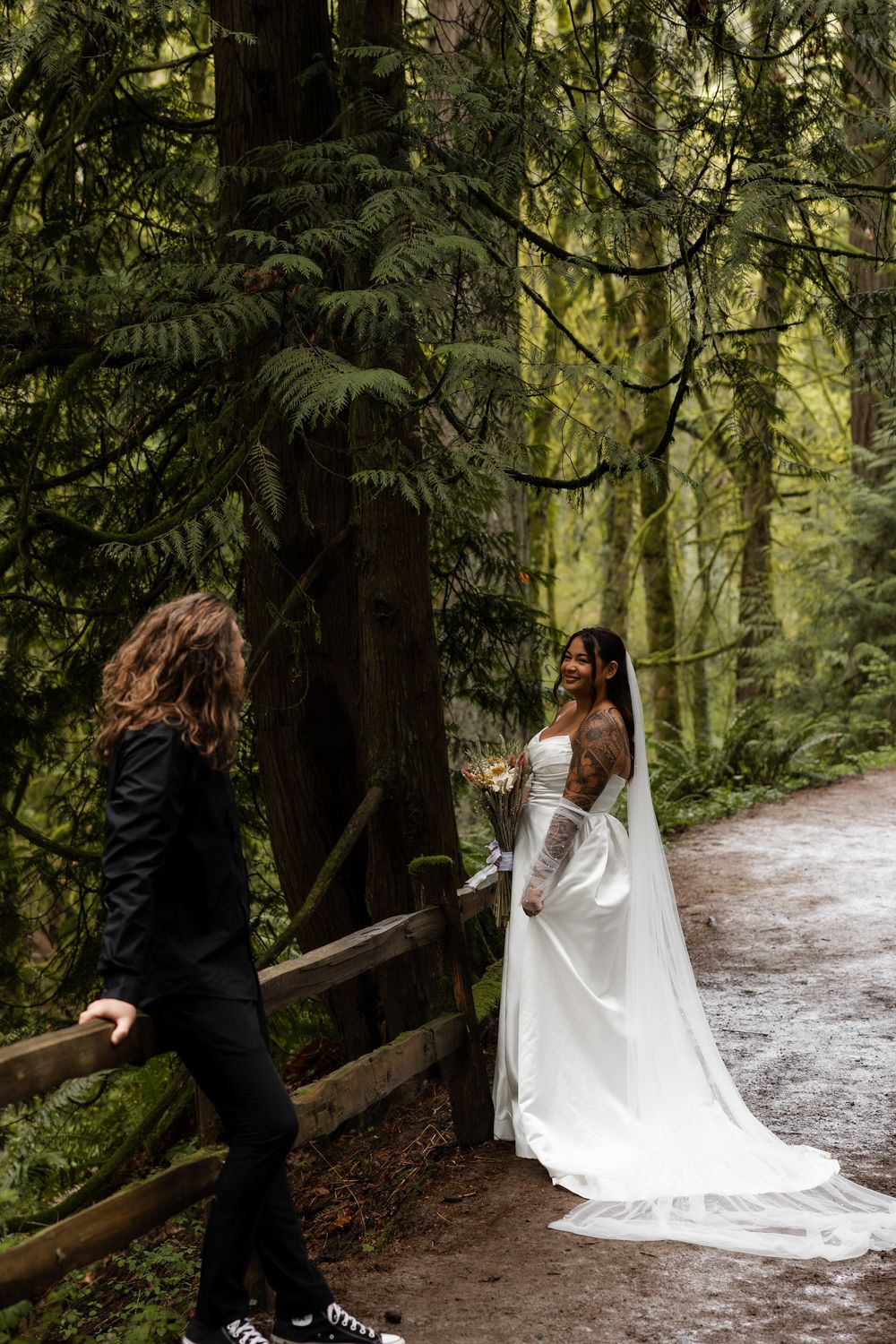 Bride in white gown with veil holds bouquet on forest path as partner in black suit approaches.