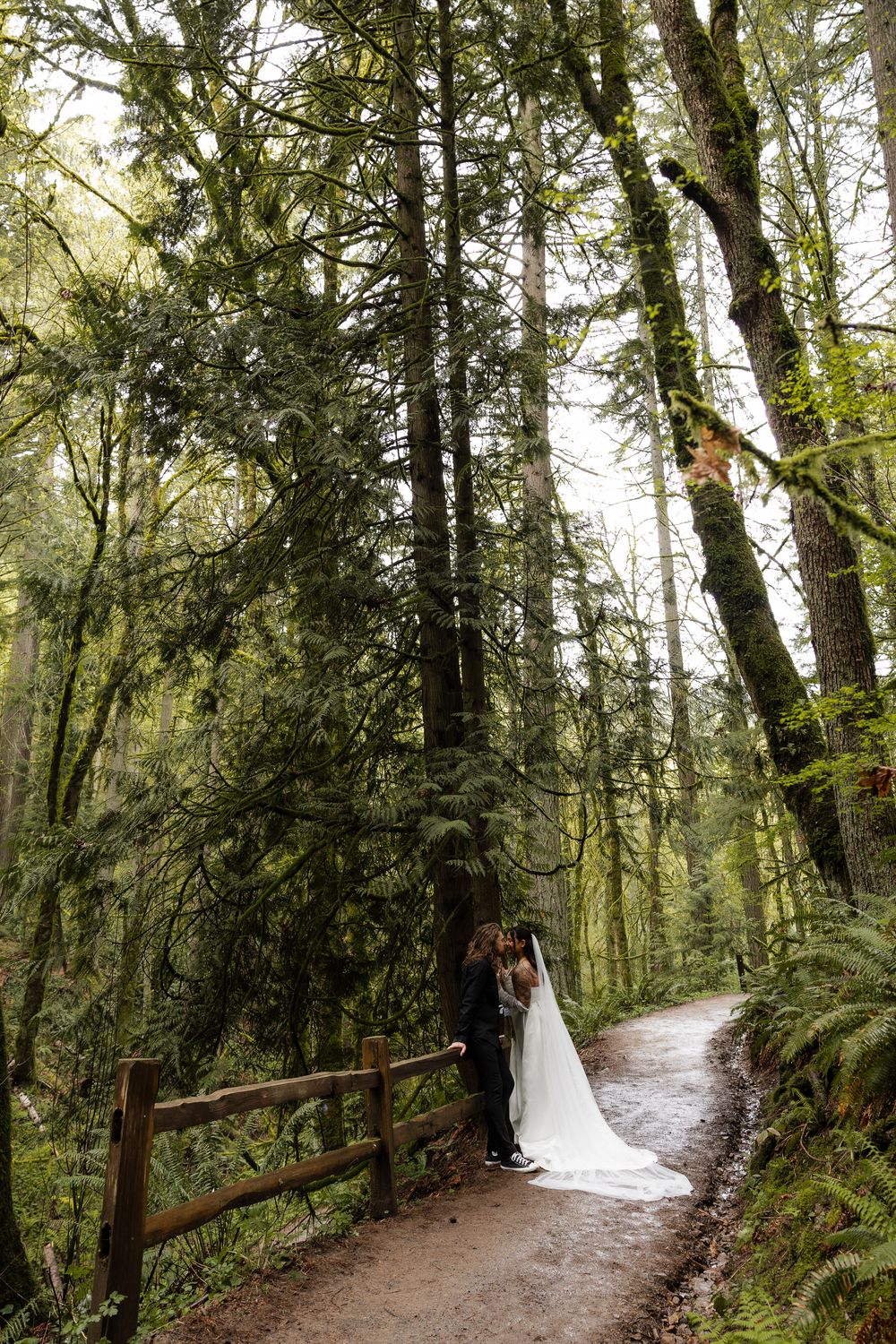 Bride and groom share a romantic embrace on a forest path surrounded by tall moss-covered trees.