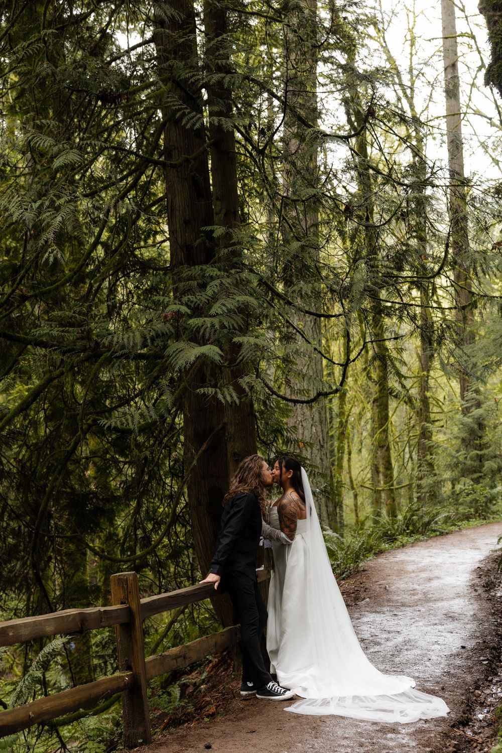 Couple shares a kiss on a mossy forest trail, bride in white gown and veil, groom in black suit.