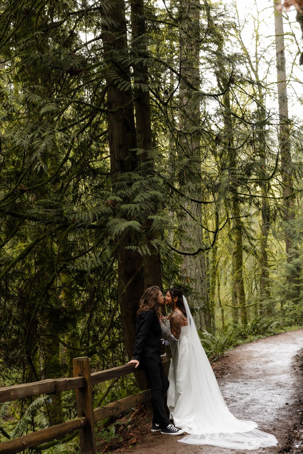 Bride and groom sharing a romantic kiss on a misty forest path beside a wooden fence.