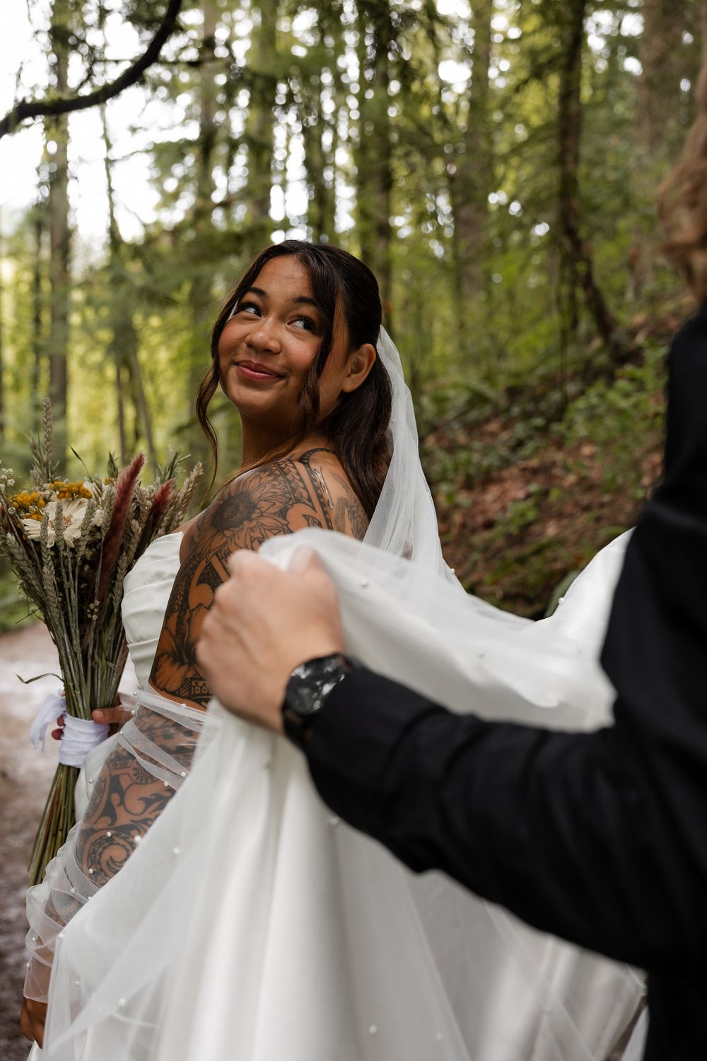 Tattooed bride in white gown smiles over shoulder as groom adjusts her veil in lush forest setting.