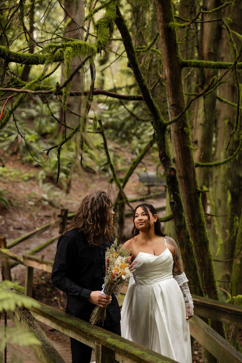 Bride and groom share a moment on a wooden bridge surrounded by mossy forest trees.