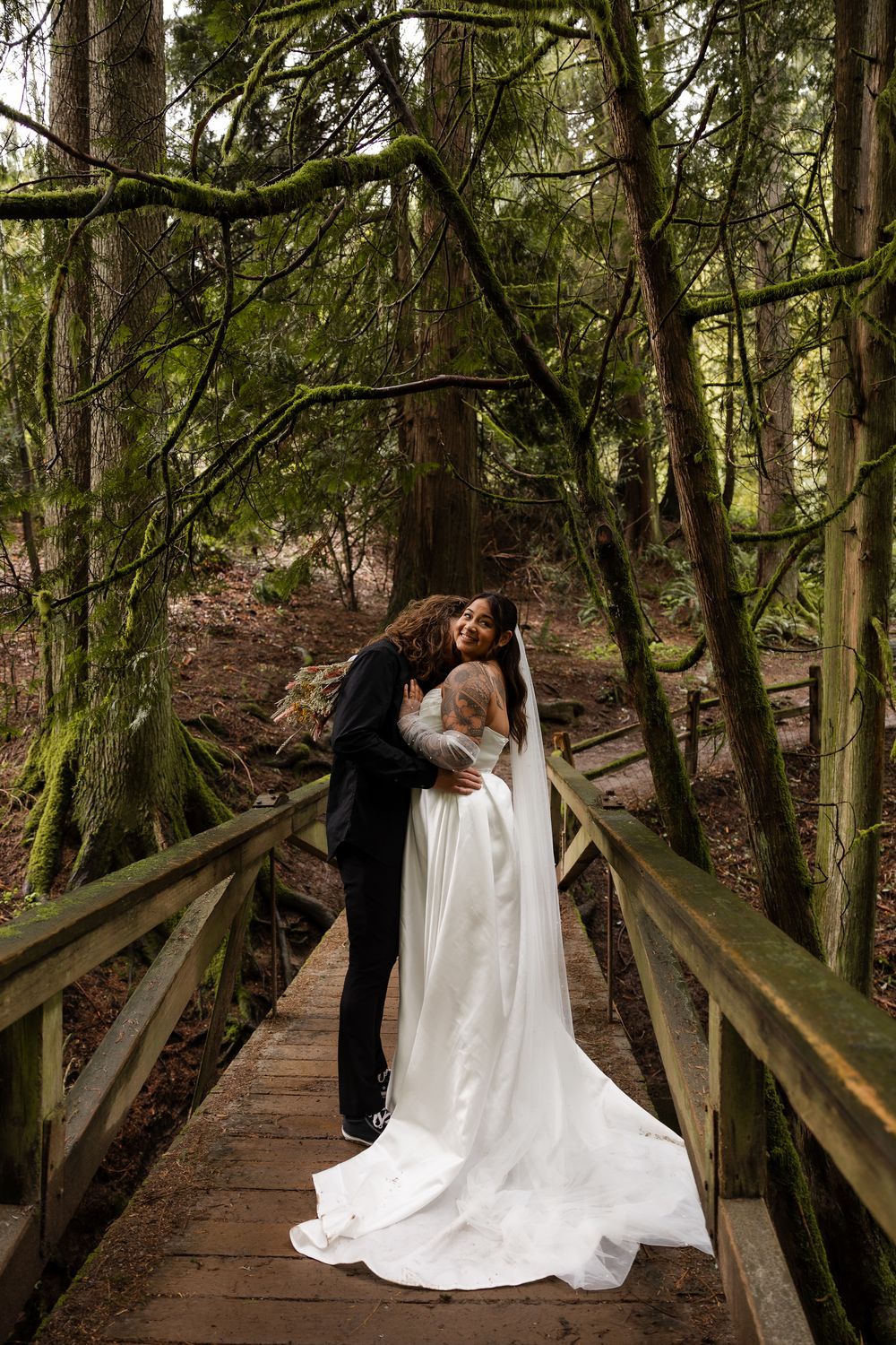 Bride and groom embrace on wooden forest bridge, she wears a flowing white gown with veil, surrounded by tall trees.